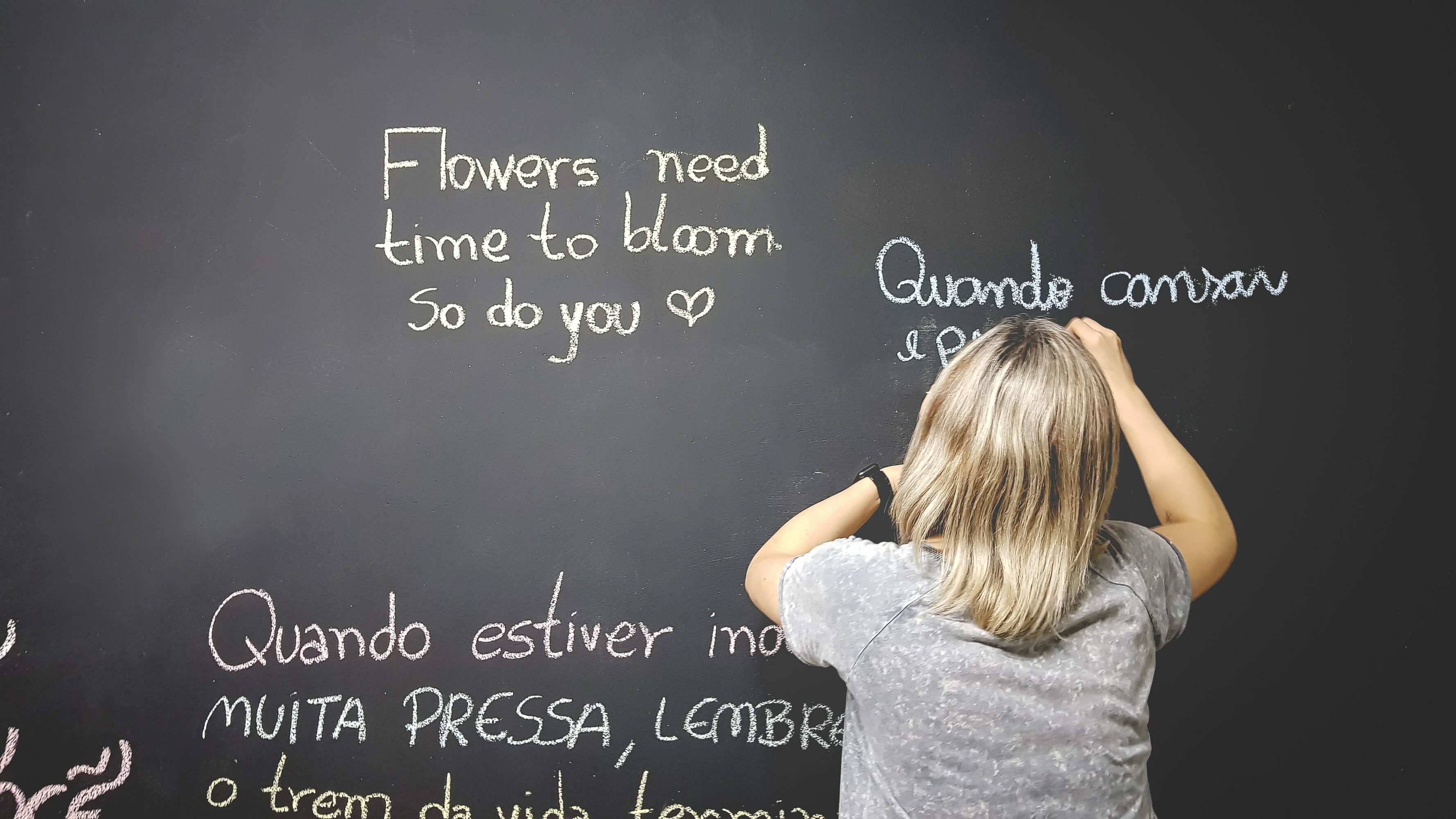 An adult writing on a chalkboard. One sentiment says "Flowers need time to bloom. So do you.