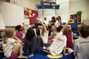 a classroom of young students sitting on the rug listening to their teacher and other students speak.
