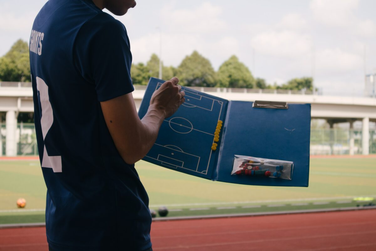 An adult standing next to a soccer field with a playbook for coaching.