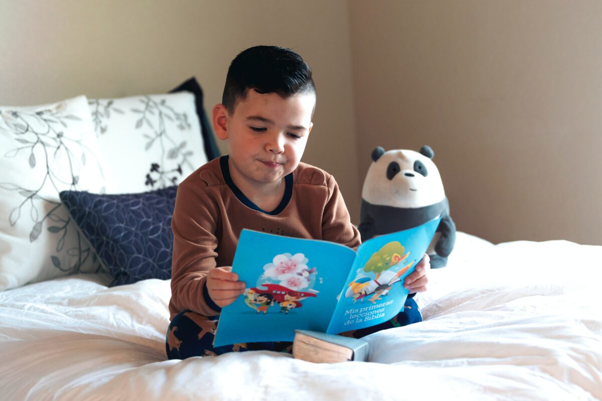 A young child sitting on a bed, reading a book with a stuffed panda bear.