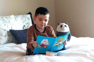 A young child sitting on a bed, reading a book with a stuffed panda bear.