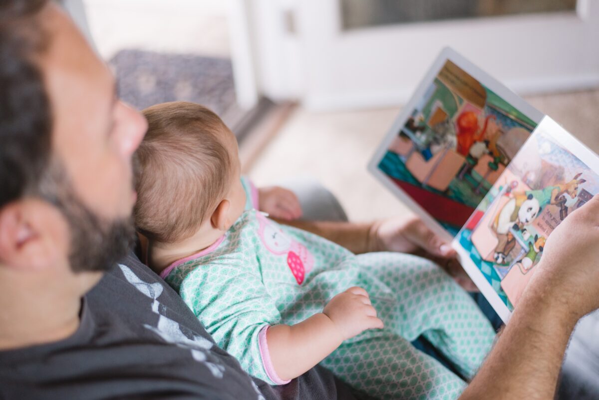 An adult is holding a baby, reading a board book.